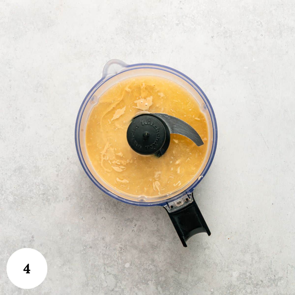 A food processor filled with a smooth, light yellow hummus mixture sits on a light gray surface. A white circle in the lower left corner displays the number 4.