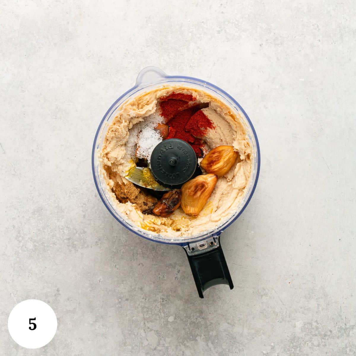 Overhead view of a food processor with creamy hummus ingredients-chickpeas, garlic cloves, spices, olive oil, and seasonings-on a light gray surface. A white circle with the number 5 is in the bottom left corner.