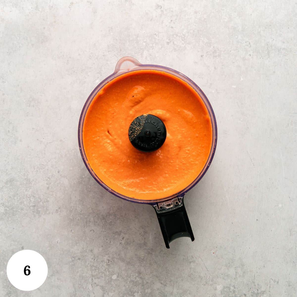 A food processor bowl filled with smooth, bright orange Roasted Red Bell Pepper Hummus sits on a light gray surface. The processor lid is open, and the number 6 is in a white circle at the bottom left corner.