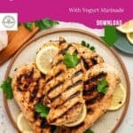 A plate of grilled Greek chicken with yogurt marinade, garnished with lemon slices and parsley, set on a table with lemon, garlic, and spices. Text above reads “Greek Chicken With Yogurt Marinade.”.