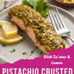 A pistachio crusted salmon fillet is topped with a vibrant green pistachio crust, garnished with parsley and lemon slices, and served on a plate with a fork. Text reads: “With Za’atar & Lemon. Pistachio Crusted Salmon.”.