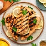 Three grilled chicken breasts with visible char marks, prepared as Greek chicken with yogurt marinade, are served on a plate with lemon slices and fresh parsley. The plate is surrounded by lemon wedges, parsley, and a garlic bulb on a light surface.