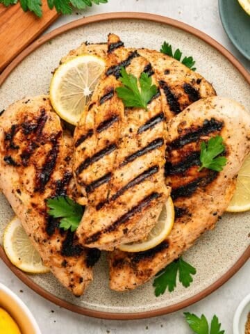 Three grilled chicken breasts with visible char marks, prepared as Greek chicken with yogurt marinade, are served on a plate with lemon slices and fresh parsley. The plate is surrounded by lemon wedges, parsley, and a garlic bulb on a light surface.