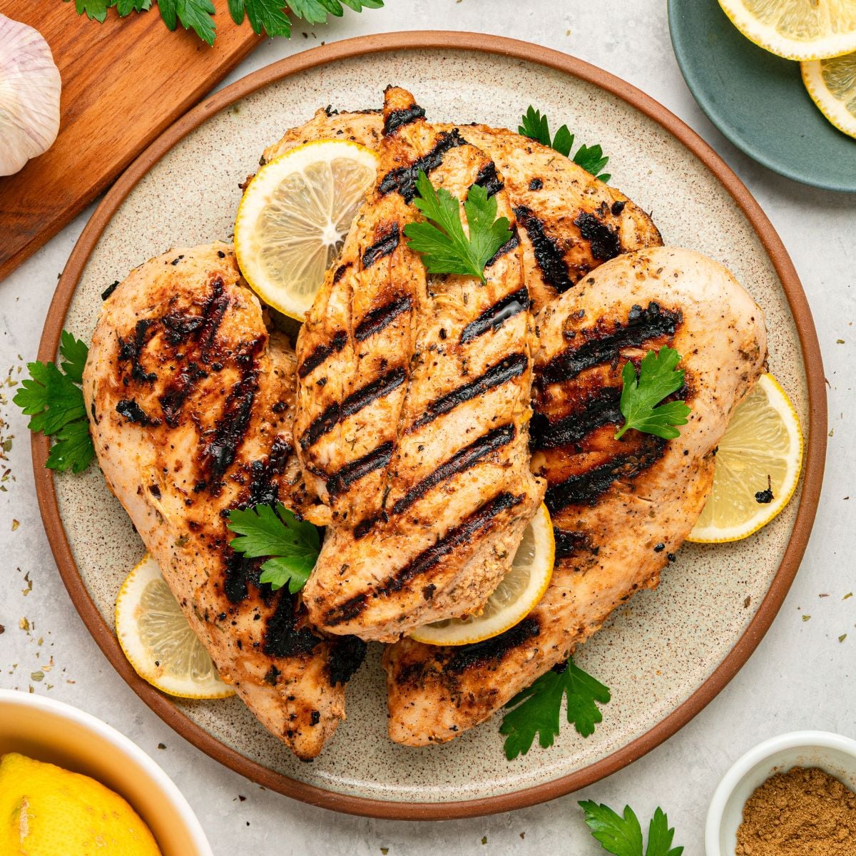 Three grilled chicken breasts with visible char marks, prepared as Greek chicken with yogurt marinade, are served on a plate with lemon slices and fresh parsley. The plate is surrounded by lemon wedges, parsley, and a garlic bulb on a light surface.