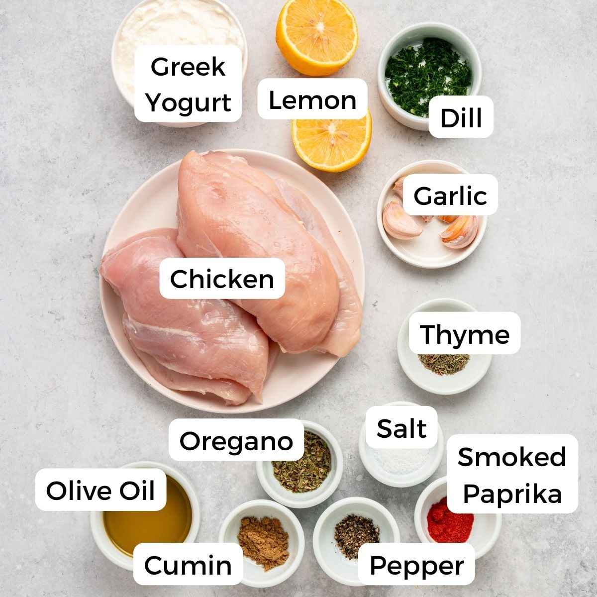 A flat lay of ingredients for greek chicken with yogurt marinade, featuring raw chicken breasts, Greek yogurt, half a lemon, chopped dill, garlic cloves, thyme, smoked paprika, salt, pepper, oregano, cumin, and olive oil in small bowls.