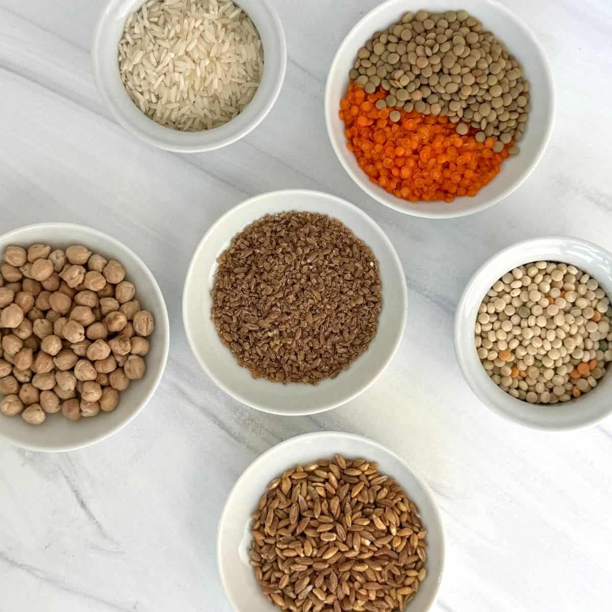 Six white bowls on a white surface, each filled with Mediterranean pantry staples—white rice, lentils, chickpeas, flax seeds, and whole grains—are arranged in a circle, showcasing a vibrant selection of dry foods.