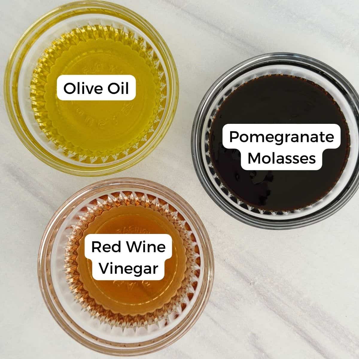 Three small glass bowls on a white surface, each containing a different liquid—olive oil, pomegranate molasses, and red wine vinegar—are labeled clearly, showcasing classic Mediterranean pantry staples.