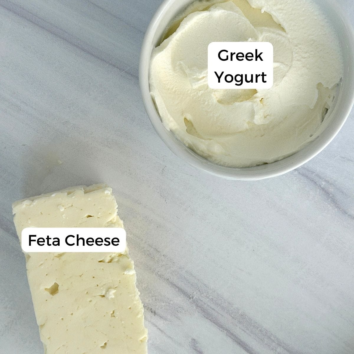 A block of feta cheese and a bowl of Greek yogurt, classic Mediterranean pantry staples, are placed on a light-colored surface. Each item is labeled with its name: Feta Cheese and Greek Yogurt.