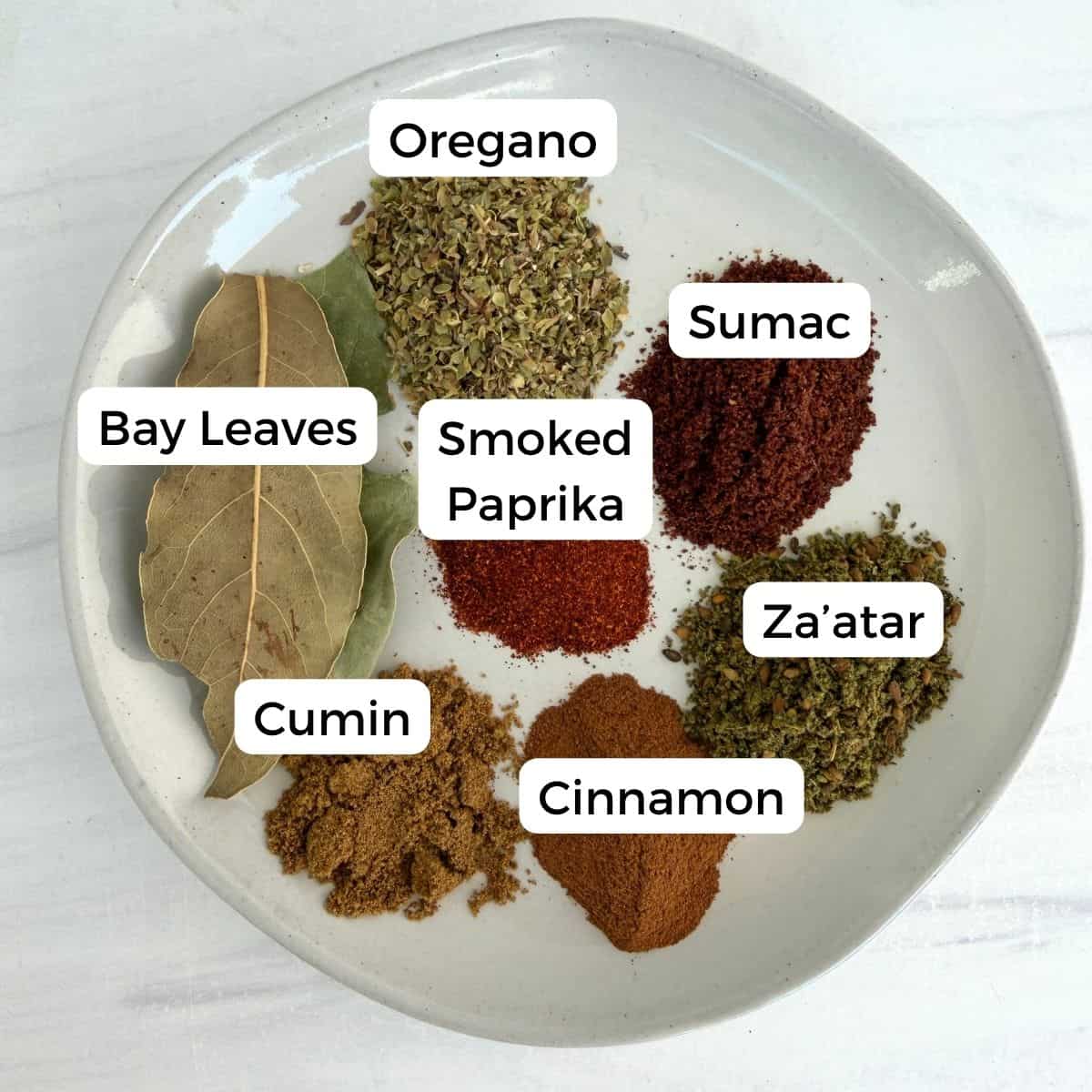 A white plate with small piles of assorted Mediterranean pantry staples—oregano, sumac, za’atar, cinnamon, cumin, smoked paprika, and two dried bay leaves. Each spice is labeled and the plate sits on a light surface.