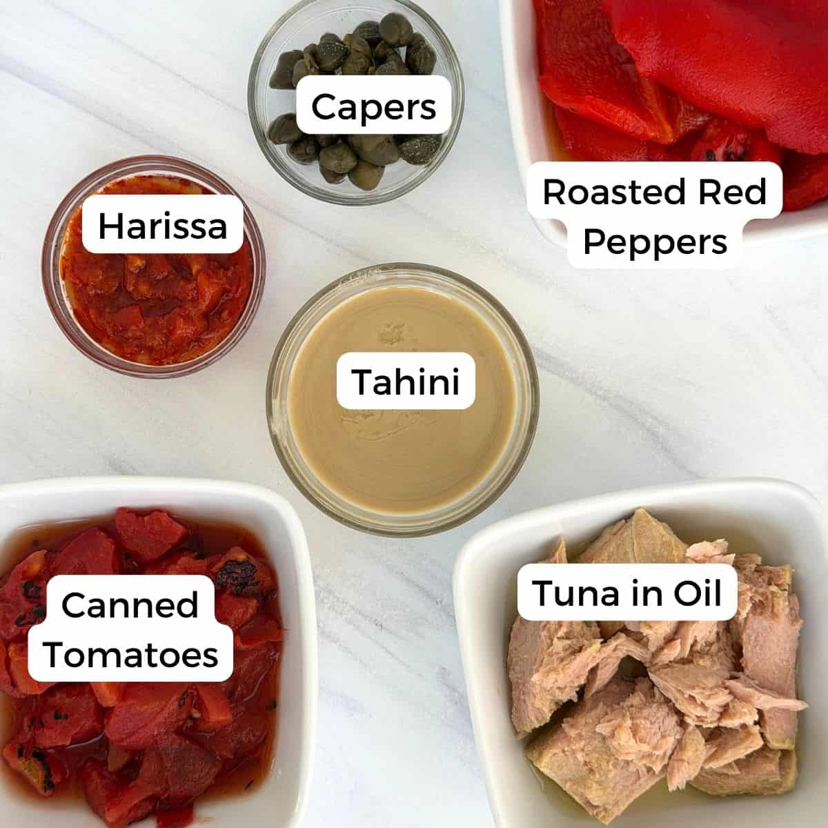 Five bowls display Mediterranean pantry staples on a white surface: capers, roasted red peppers, harissa, tahini, canned tomatoes, and tuna in oil. Each ingredient is labeled with text.