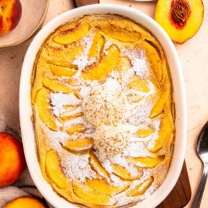 A baked peach cobbler in a white dish, reminiscent of a peach clafoutis, is topped with two scoops of vanilla ice cream and sprinkled with powdered sugar. Sliced peaches and peach halves are visible around the dish.