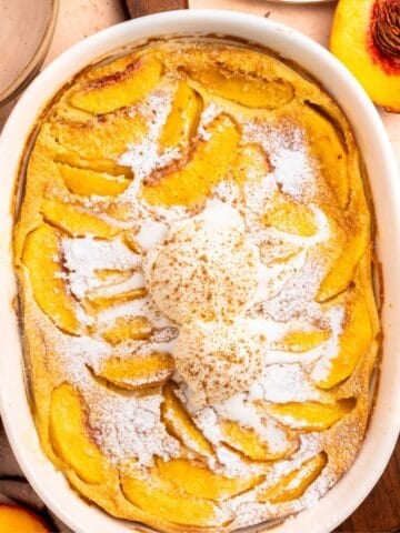 A baked peach cobbler in a white dish, reminiscent of a peach clafoutis, is topped with two scoops of vanilla ice cream and sprinkled with powdered sugar. Sliced peaches and peach halves are visible around the dish.
