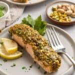 A plate with a slice of pistachio crusted salmon, topped with a vibrant green pistachio crust and garnished with lemon slices and parsley. A fork rests on the plate, with a dish of pistachios in the background.