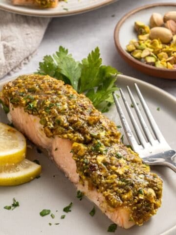 A plate with a slice of pistachio crusted salmon, topped with a vibrant green pistachio crust and garnished with lemon slices and parsley. A fork rests on the plate, with a dish of pistachios in the background.