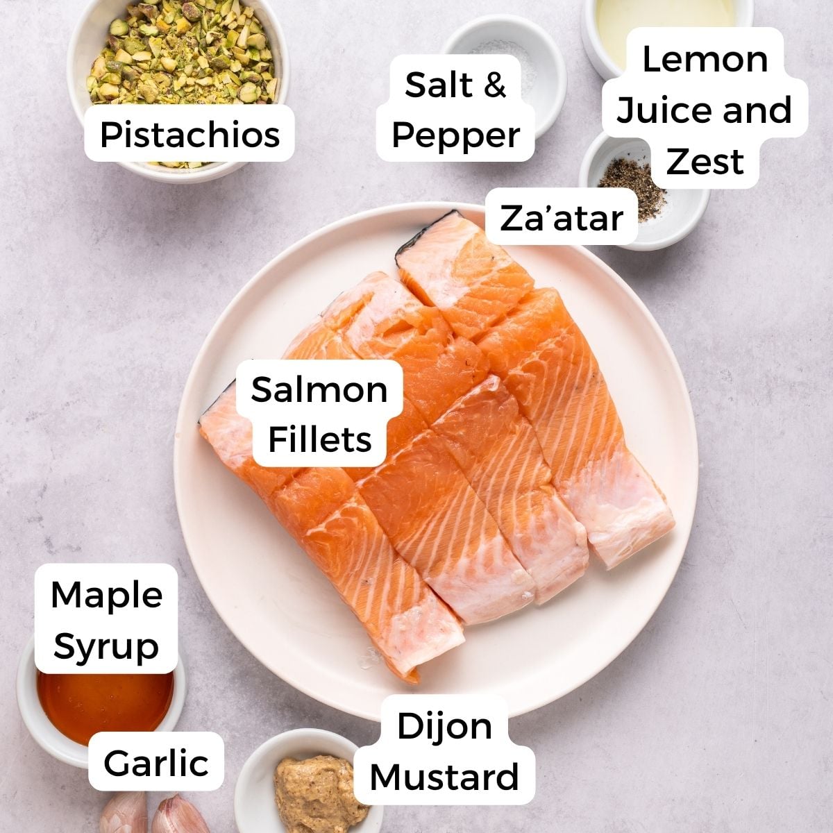 Top-down view of a plate with raw salmon fillets surrounded by labeled ingredients for pistachio crusted salmon: pistachios, salt & pepper, lemon juice and zest, za’atar, maple syrup, garlic, and Dijon mustard on a light surface.