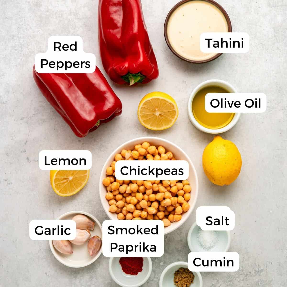 Top-down view of ingredients for Creamy Hummus, including red peppers, tahini, olive oil, lemon, chickpeas, garlic, smoked paprika, salt, and cumin—all labeled with white text on a light background.