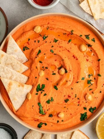 A bowl of creamy roasted red bell pepper hummus garnished with chickpeas, parsley, and paprika, surrounded by pita chips and lemon halves on a light surface.