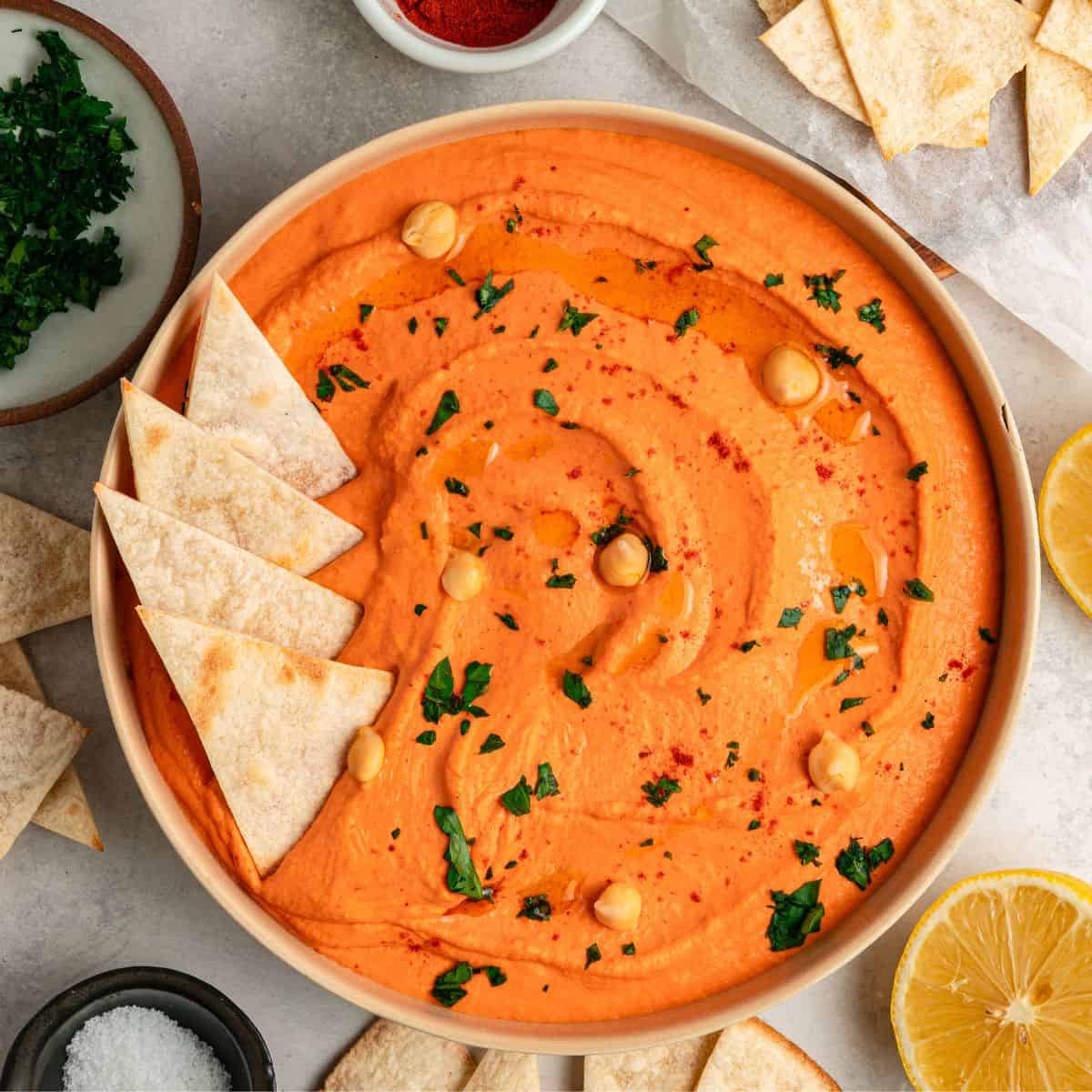 A bowl of creamy roasted red bell pepper hummus garnished with chickpeas, parsley, and paprika, surrounded by pita chips and lemon halves on a light surface.