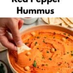 A hand dips a pita chip into a bowl of vibrant Roasted Red Bell Pepper Hummus topped with chickpeas and herbs. Text above reads Smokey & Creamy Red Pepper Hummus. Tortilla chips and a dip are visible in the background.