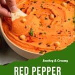 A hand dips a tortilla chip into a bowl of Roasted Red Bell Pepper Hummus, garnished with chickpeas, herbs, and paprika. Text on image reads “Smokey & Creamy Red Pepper Hummus.”.