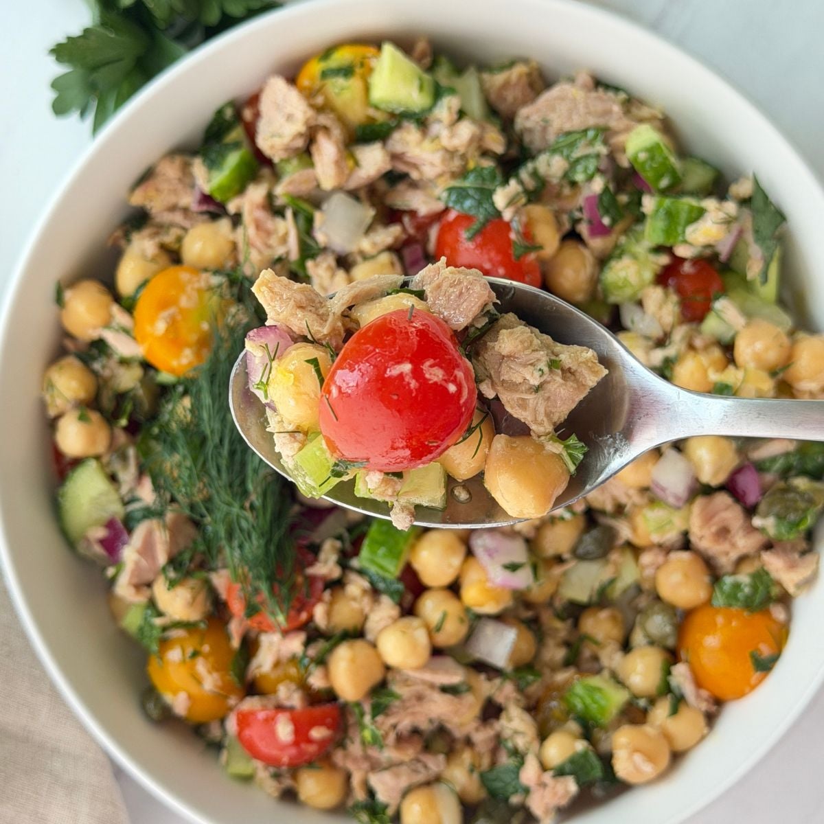 A spoonful of chickpea tuna salad with cherry tomatoes, cucumber, red onion, herbs, and parsley is held above a bowl containing the same colorful salad.