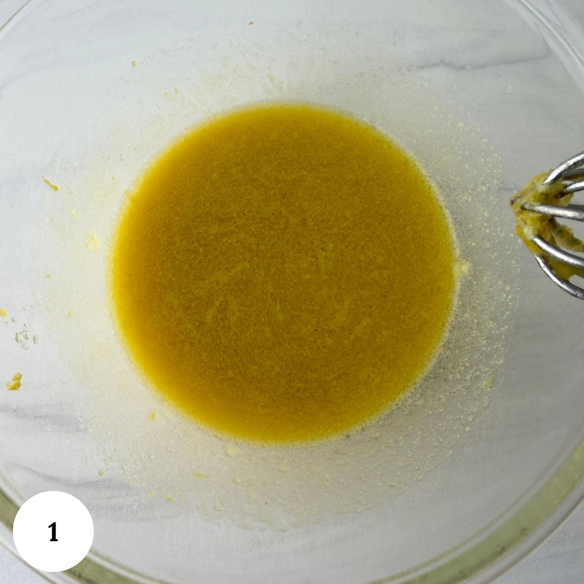 A glass mixing bowl with a yellowish liquid mixture and a metal whisk, seen from above-perfect for prepping ingredients like those in a chickpea tuna salad. The number 1 is in a white circle in the bottom left corner.