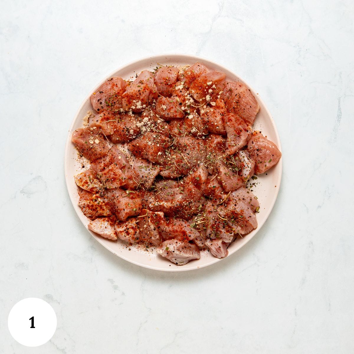 Raw chicken pieces on a white plate, perfectly seasoned with various spices and herbs for garlic parmesan pasta with chicken, set on a light marble surface. A white circle with the number 1 is in the bottom left corner.