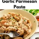 A bowl of creamy garlic parmesan pasta with chicken, garnished with herbs, sits on a table alongside fresh parsley, aromatic spices, and a fork. Text on the image reads “Garlic Parmesan Pasta With Chicken,” highlighting this delicious and comforting meal.