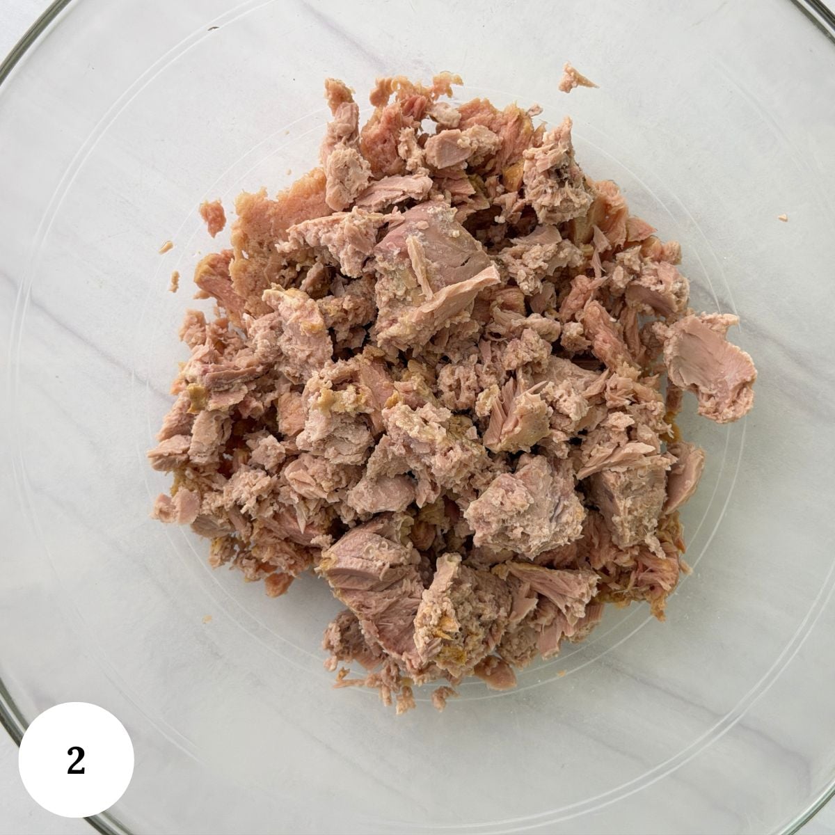 A clear glass bowl containing flaked, light brown canned tuna for a chickpea tuna salad is placed on a white surface. A white circle with the number 2 is in the lower left corner of the image.