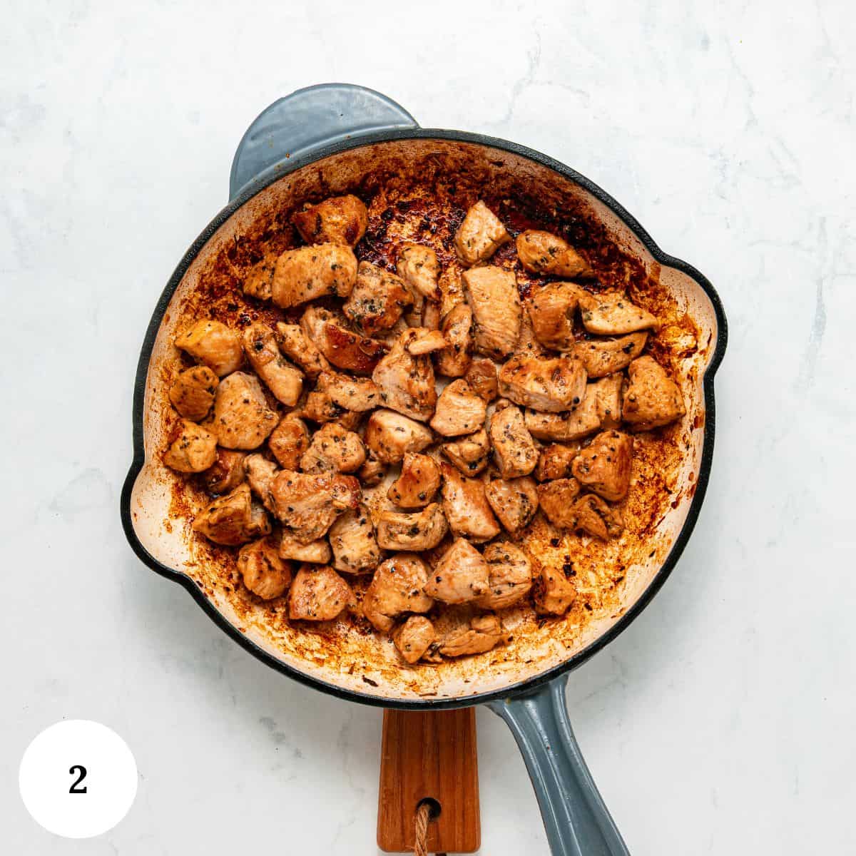 A skillet filled with browned, seasoned chicken pieces-perfectly cooked for a garlic parmesan pasta with chicken-rests on a light surface. A small white circle with the number 2 is in the bottom left corner of the image.