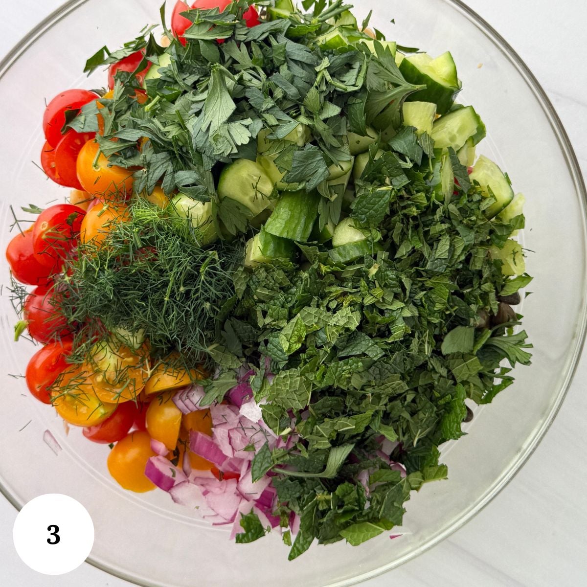 A glass bowl filled with chopped cherry tomatoes, cucumber, red onion, parsley, dill, and mint sits ready to be mixed into a fresh chickpea tuna salad. A white circle with the number 3 is in the bottom left corner.