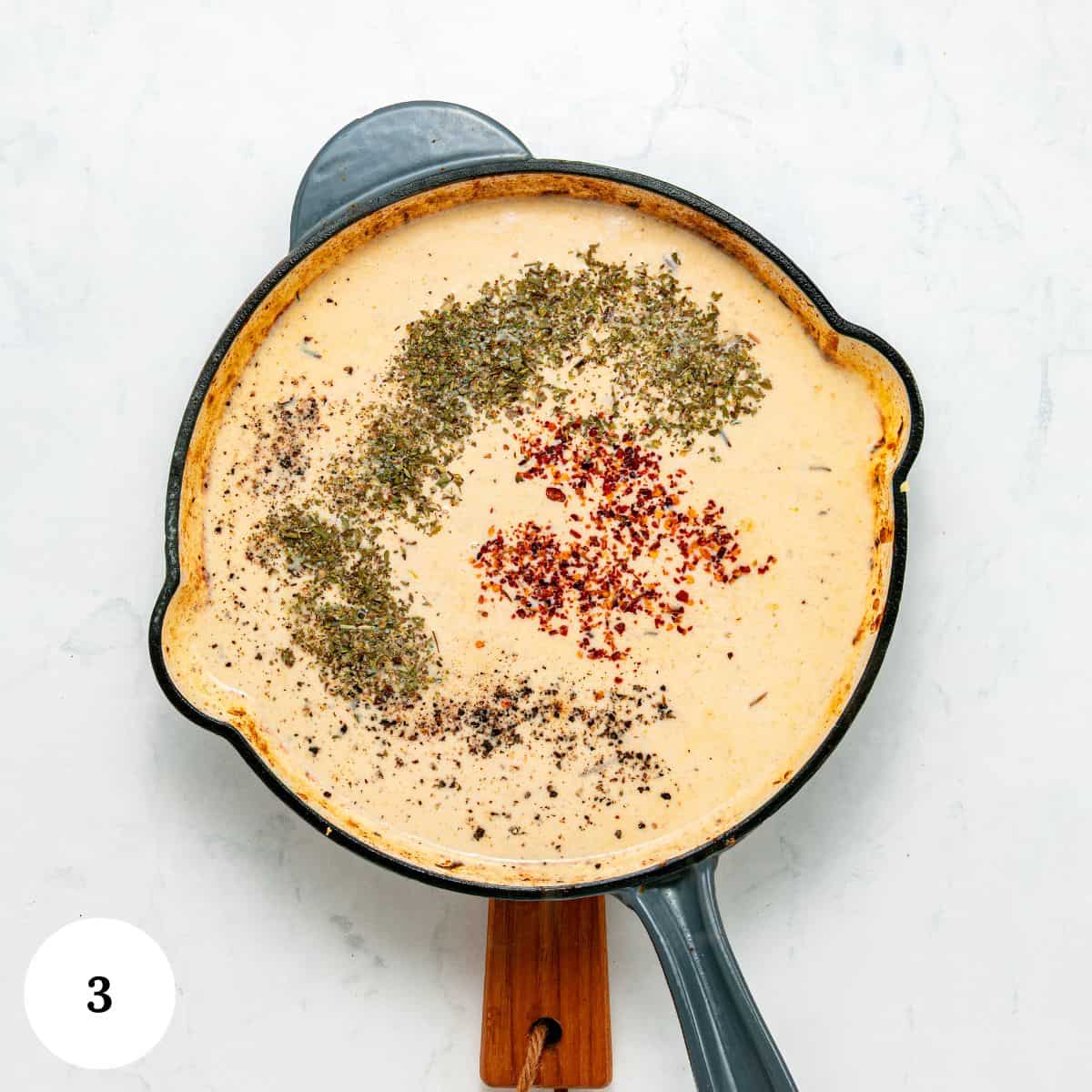 A skillet of creamy garlic parmesan pasta with chicken, topped with dried herbs, red pepper flakes, and black pepper, sits on a wooden handle, viewed from above on a white surface. A circle with the number 3 is in the bottom left corner.