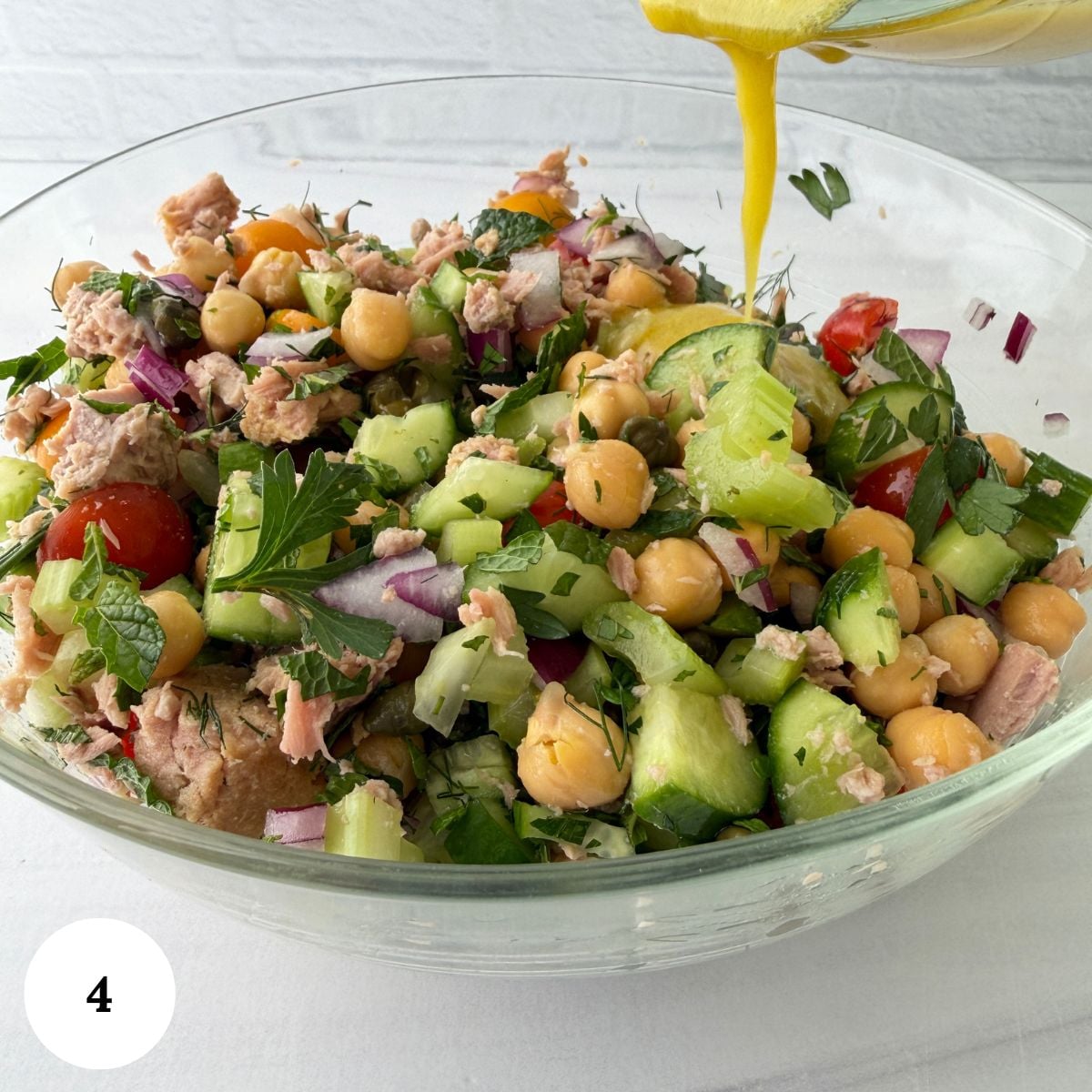 A glass bowl filled with a vibrant chickpea tuna salad-tuna, chickpeas, cucumber, tomatoes, red onion, and parsley-all brought together as yellow dressing is poured over the fresh ingredients.