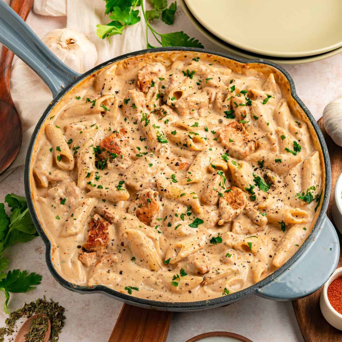 A skillet filled with creamy garlic parmesan pasta with chicken pieces, garnished with chopped parsley and black pepper. Garlic, herbs, and empty plates are arranged around the dish on a light-colored surface.