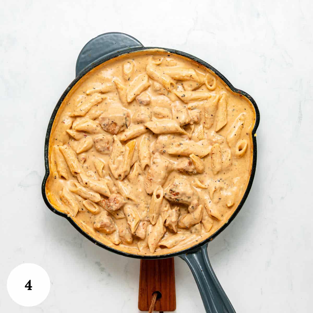 A skillet filled with creamy garlic parmesan pasta with chicken-tender penne and juicy pieces of chicken in a rich, orange-tinted sauce-sits on a white surface. A wooden handle extends from the pan. A circular label with the number 4 is in the lower left corner.