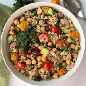 Bowl of vibrant chickpea tuna salad with cherry tomatoes, cucumber, red onion, parsley, and dill, placed on a white surface with a green napkin, spoon, and fresh parsley nearby.