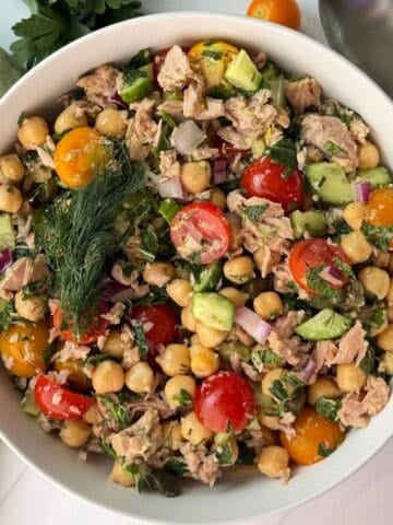 Bowl of vibrant chickpea tuna salad with cherry tomatoes, cucumber, red onion, parsley, and dill, placed on a white surface with a green napkin, spoon, and fresh parsley nearby.