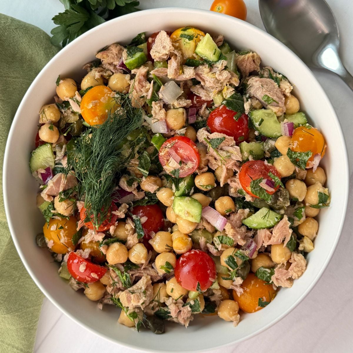 Bowl of vibrant chickpea tuna salad with cherry tomatoes, cucumber, red onion, parsley, and dill, placed on a white surface with a green napkin, spoon, and fresh parsley nearby.