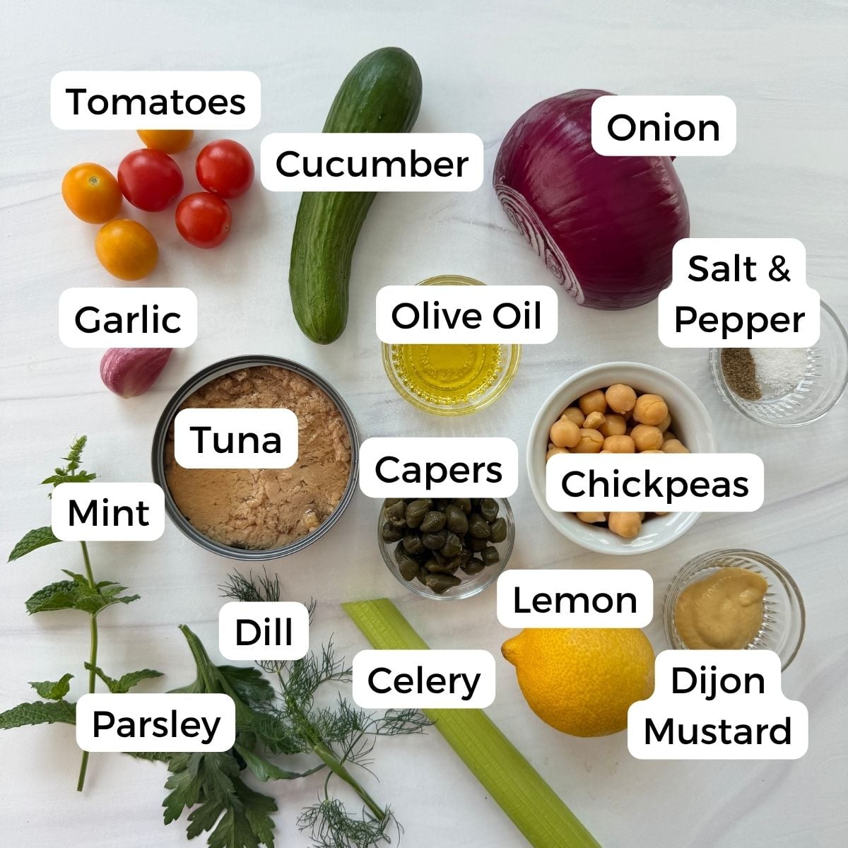A variety of labeled ingredients for a chickpea tuna salad are displayed on a white surface, including tomatoes, cucumber, onion, garlic, tuna, olive oil, salt, pepper, chickpeas, capers, lemon, Dijon mustard, celery, dill, mint, and parsley.