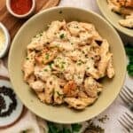 A bowl of creamy garlic parmesan pasta with chicken, topped with tender grilled pieces, is sprinkled with chopped parsley and parmesan cheese. Surrounding the bowl are peppercorns, grated cheese, forks, and a small bowl of paprika.