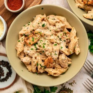 A bowl of creamy garlic parmesan pasta with chicken, topped with tender grilled pieces, is sprinkled with chopped parsley and parmesan cheese. Surrounding the bowl are peppercorns, grated cheese, forks, and a small bowl of paprika.