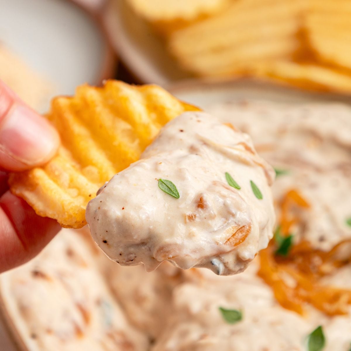 A hand holds a crinkle-cut potato chip dipped in creamy Greek yogurt French onion dip, garnished with small green herbs. More chips and dip are visible in the blurred background.