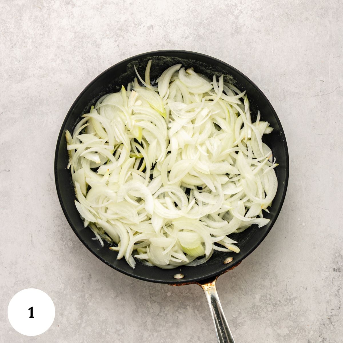 Sliced onions are spread evenly in a black frying pan on a light gray surface, ready to be slowly caramelized as the first step in making a delicious greek yogurt french onion dip. A white circle with the number 1 is at the bottom left corner.