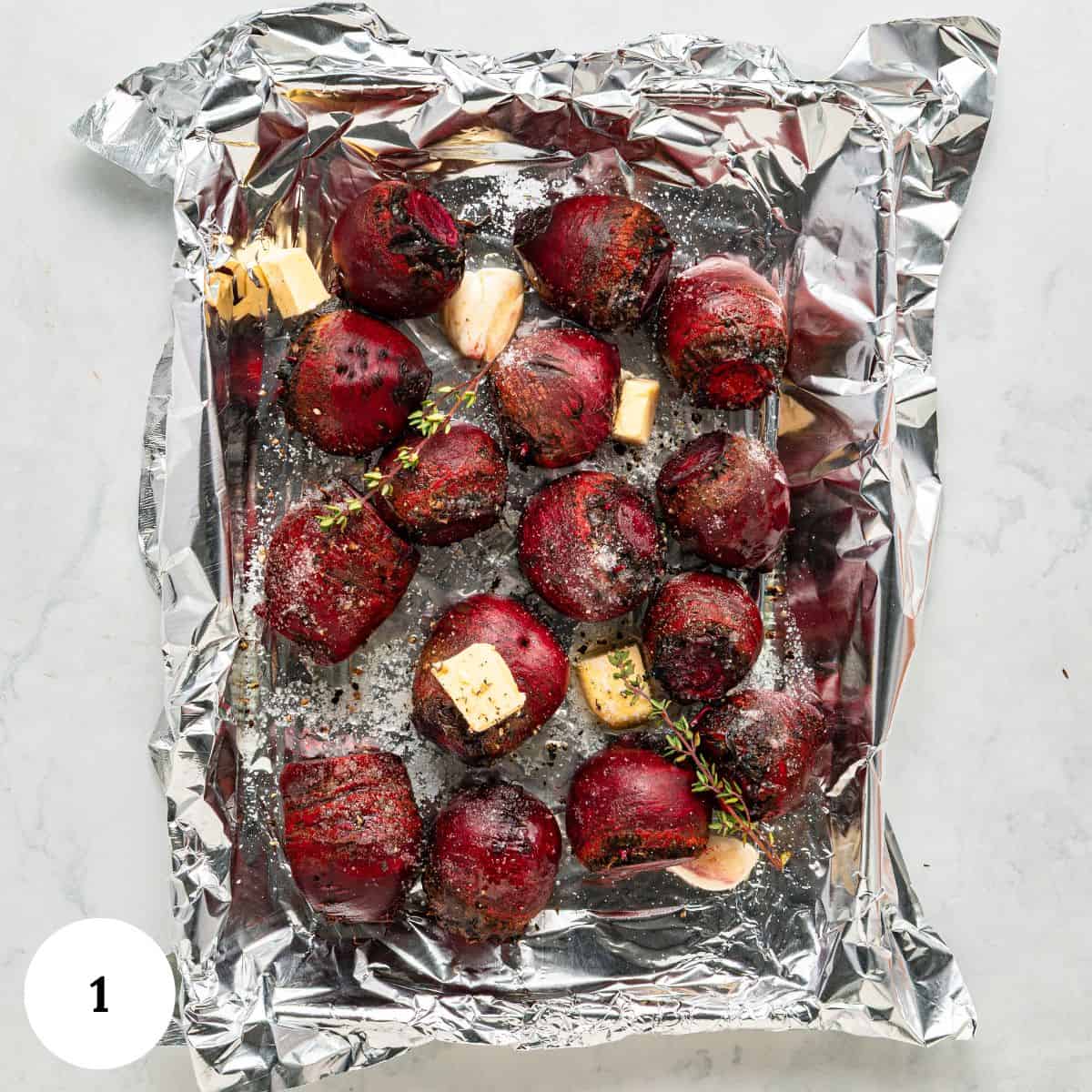 Roasted beets chunks with butter, garlic cloves, and herbs are arranged on a foil-lined baking dish, seasoned with pepper and salt, ready for roasting.