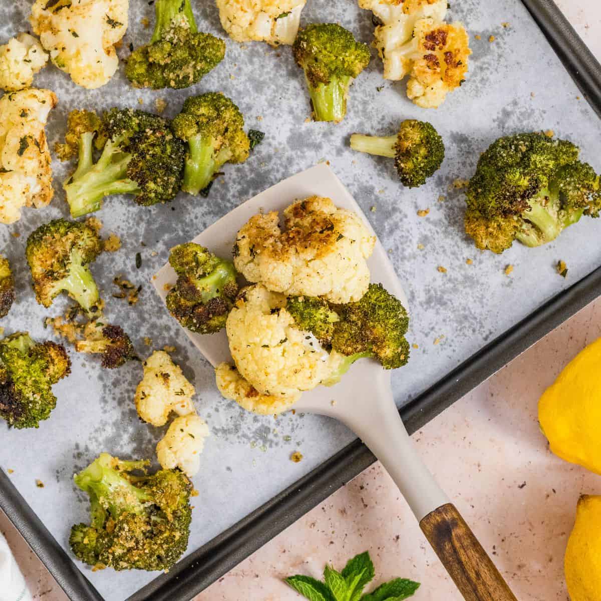 Roasted broccoli and cauliflower pieces rest on a parchment-lined baking sheet, with a spatula lifting some perfectly roasted florets. Lemons and mint leaves add a fresh touch nearby on a light-colored surface.