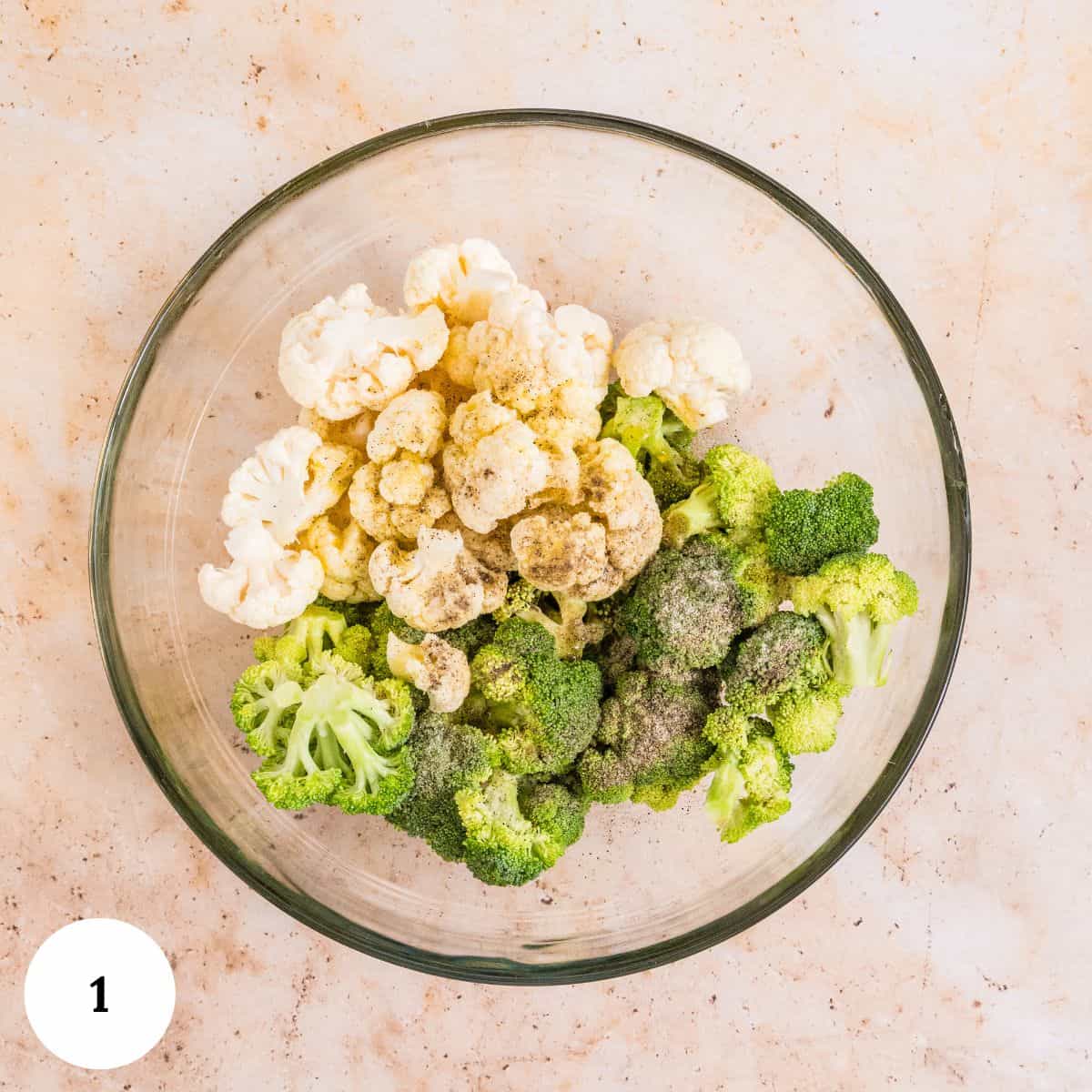 A glass bowl with raw cauliflower and broccoli florets, perfect for making roasted broccoli and cauliflower, is sprinkled with spices and sits on a light-colored countertop. A white circle with the number 1 is in the bottom left corner.