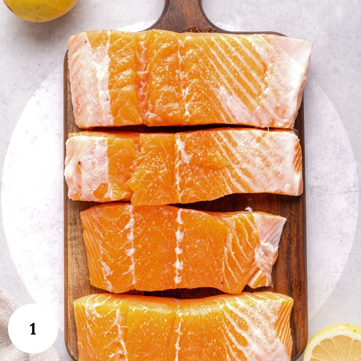 Four raw salmon fillets with white marbling are arranged on a wooden cutting board, ready to be transformed into a flavorful salmon piccata. Slices of lemon are partially visible in the corners of the image.