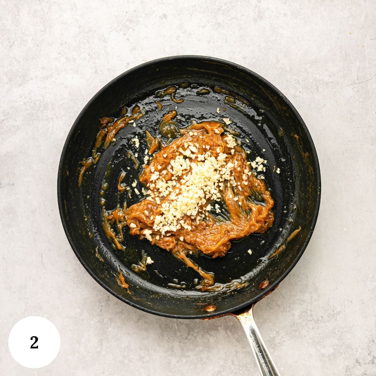 A black pan with a brown sauce and chopped garlic on top, reminiscent of the rich flavors found in greek yogurt french onion dip, sits on a light gray surface. A white circle with the number 2 is in the lower left corner.