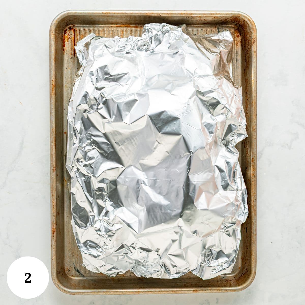 A baking sheet with roasted beets covered in crinkled aluminum foil, resting on a light surface. A small white circle with the number 2 is in the bottom left corner.