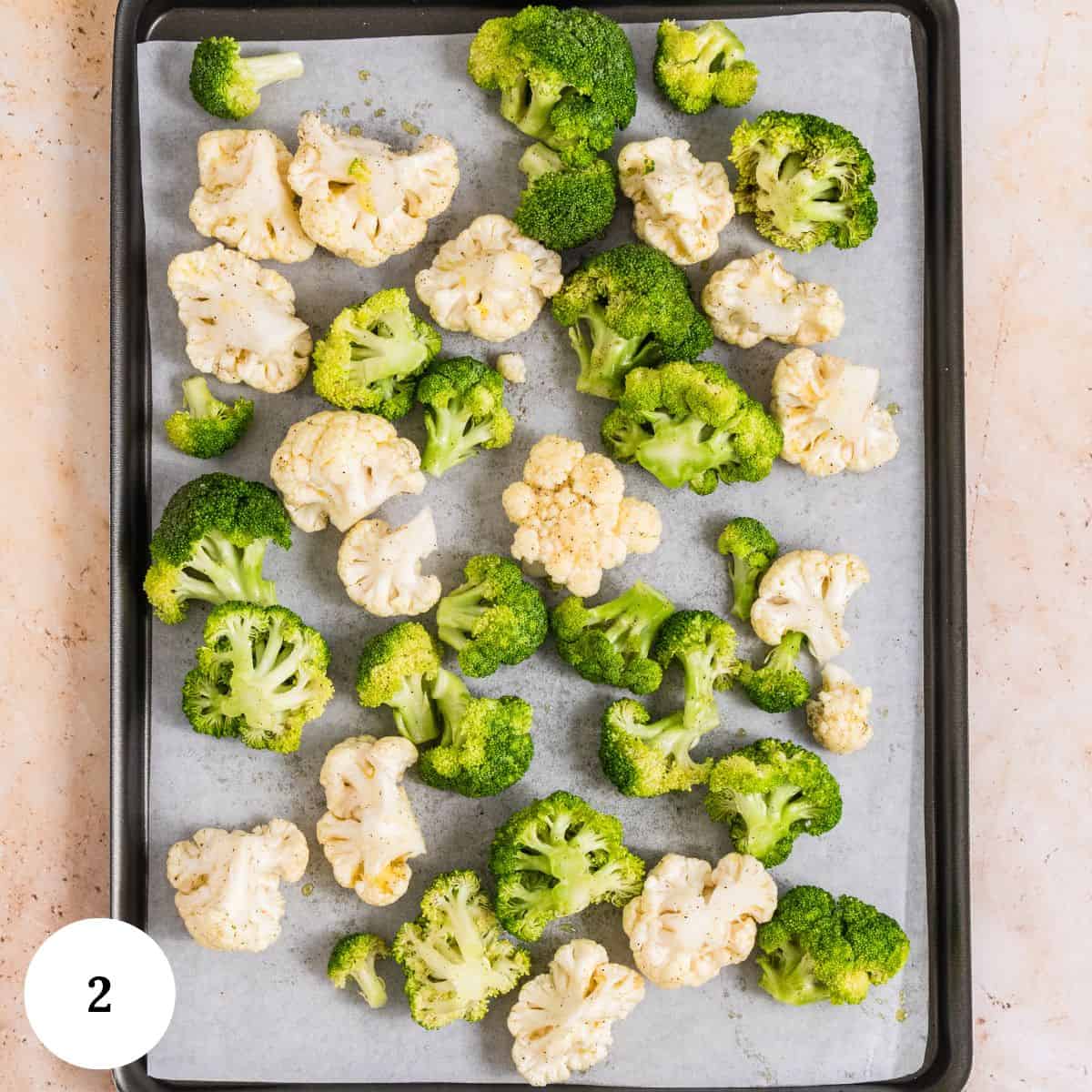 A baking sheet lined with parchment paper holds scattered raw cauliflower and broccoli florets, ready to become roasted broccoli and cauliflower. A white circle with the number 2 is in the bottom left corner.